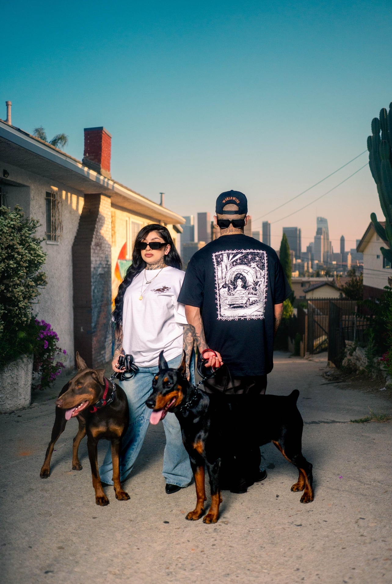 Two models standing the street with city skyline behind wear brown n proud shirts, along side two doberman dogs.